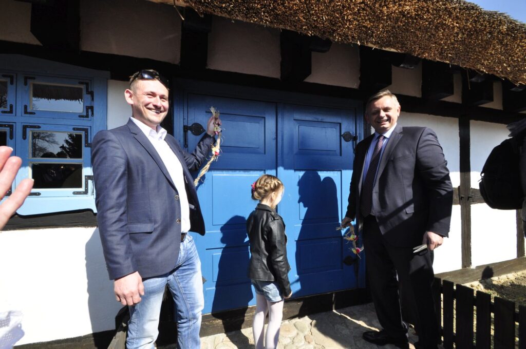 Dwóch mężczyzn w garniturach i dziecko stoją przy niebieskich drzwiach ryglowego budynku podczas symbolicznego otwarcia obiektu (kliknięcie powiększy zdjęcie).