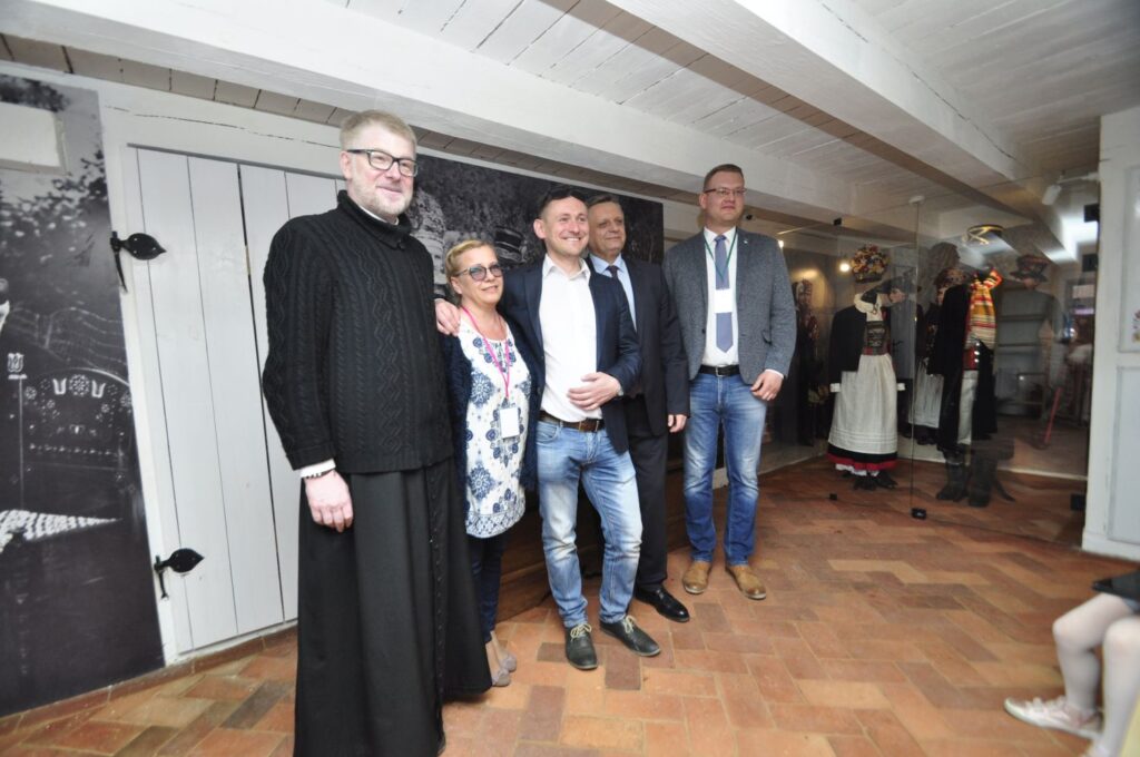 Pamiątkowe zdjęcie grupy osób, w tym duchownego, we wnętrzu chaty z białą drewnianą ścianą (kliknięcie powiększy zdjęcie).