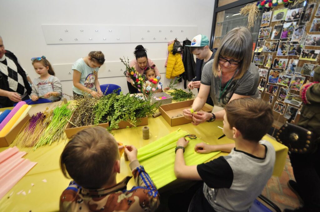 Uczestnicy warsztatów siedzą przy stole i wspólnie wykonują tradycyjne ozdoby oraz palmy z kolorowego papieru i zielonych gałązek (kliknięcie powiększy zdjęcie).