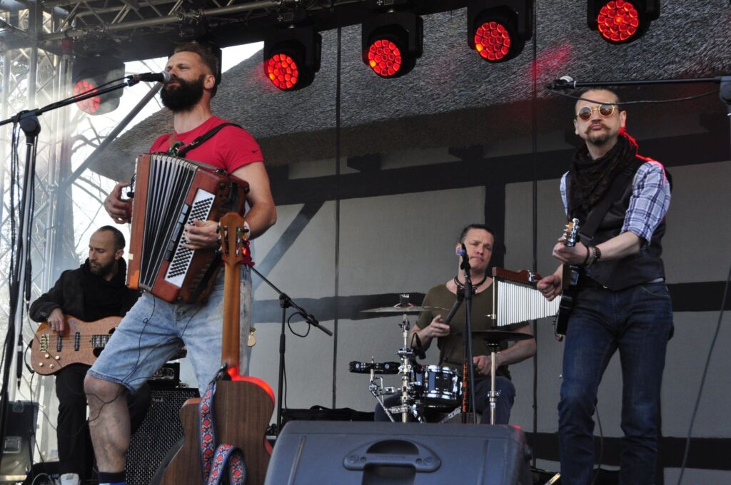 Występ zespołu muzycznego na żywo; na pierwszym planie wokalista grający na akordeonie, w tle gitarzysta i perkusista (kliknięcie powiększy zdjęcie).