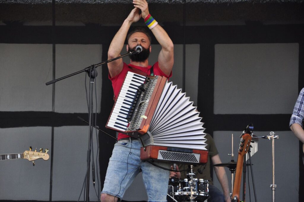 Mężczyzna w czerwonej koszulce gra na akordeonie i śpiewa do mikrofonu na scenie podczas występów plenerowych (kliknięcie powiększy zdjęcie).