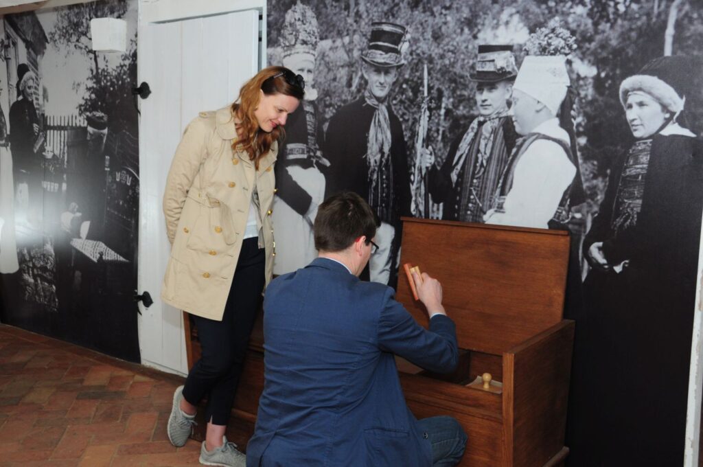 Zwiedzający oglądają ekspozycję, mężczyzna wyjmuje coś z drewnianej skrzyni, na ścianie wisi archiwalna fotografia (kliknięcie powiększy zdjęcie).