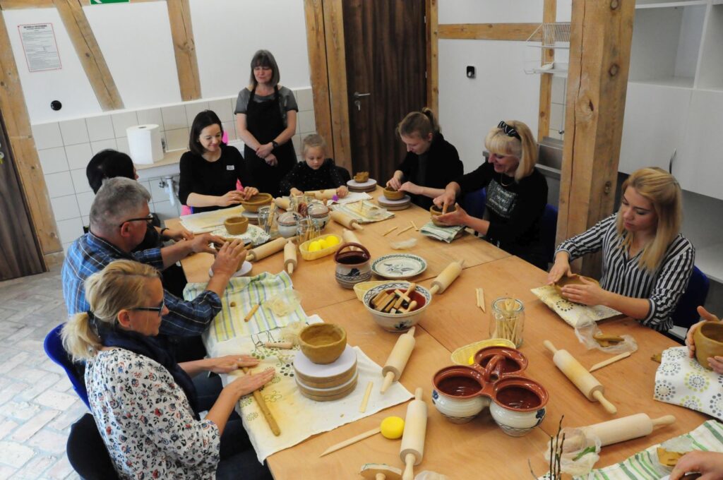 Grupa uczestników warsztatów ceramicznych siedzących wokół stołu i lepiących gliniane naczynia (kliknięcie powiększy zdjęcie).