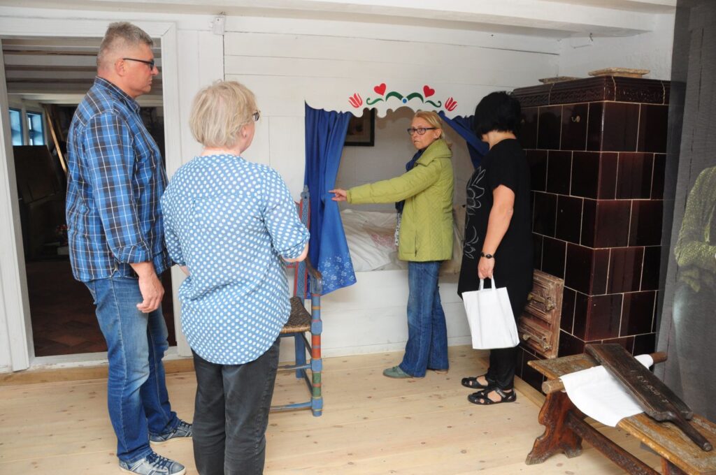 Zwiedzający oglądają rekonstrukcję dawnego wnętrza mieszkalnego z piecem kaflowym i łóżkiem, słuchając opowieści przewodnika (kliknięcie powiększy zdjęcie).