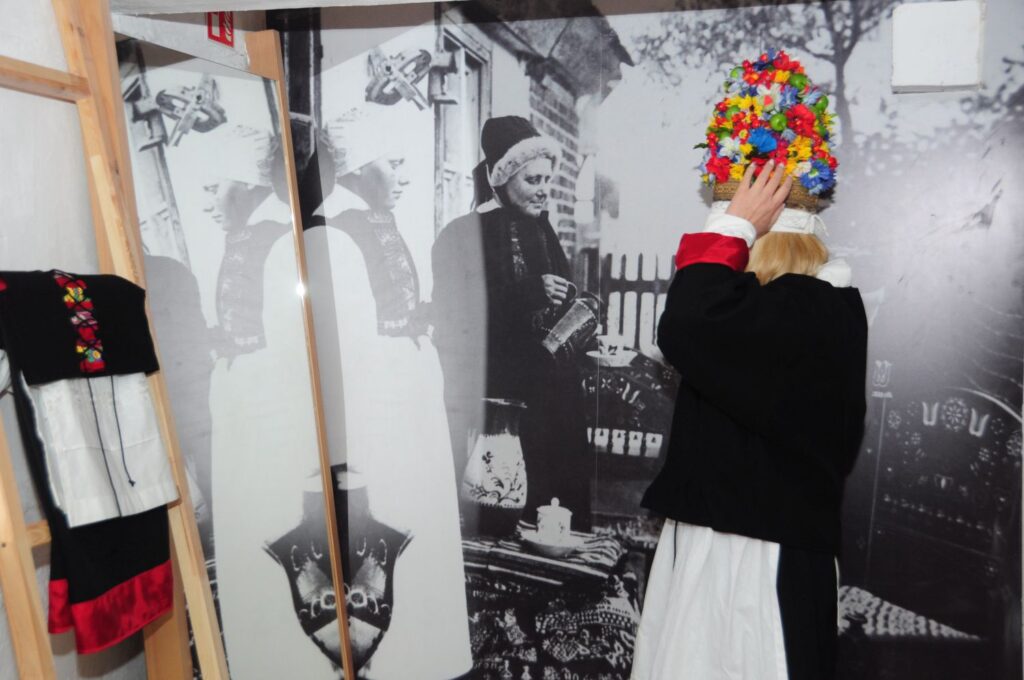 Zwiedzająca przymierza kolorowy wianek ludowy na tle archiwalnej fotografii przedstawiającej dawnych mieszkańców Jamna (kliknięcie powiększy zdjęcie).