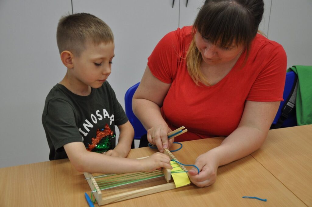 Chłopiec wykonuje tkanie na krosnach z pomocą instruktorki podczas warsztatów tkackich dla dzieci (kliknięcie powiększy zdjęcie).