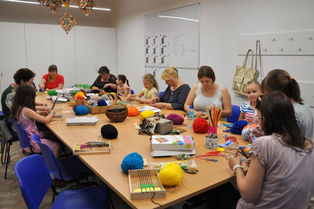 Uczestnicy warsztatów tkackich siedzą przy długim stole, pracując na krosnach i z kolorową włóczką w sali warsztatowej (kliknięcie powiększy zdjęcie).