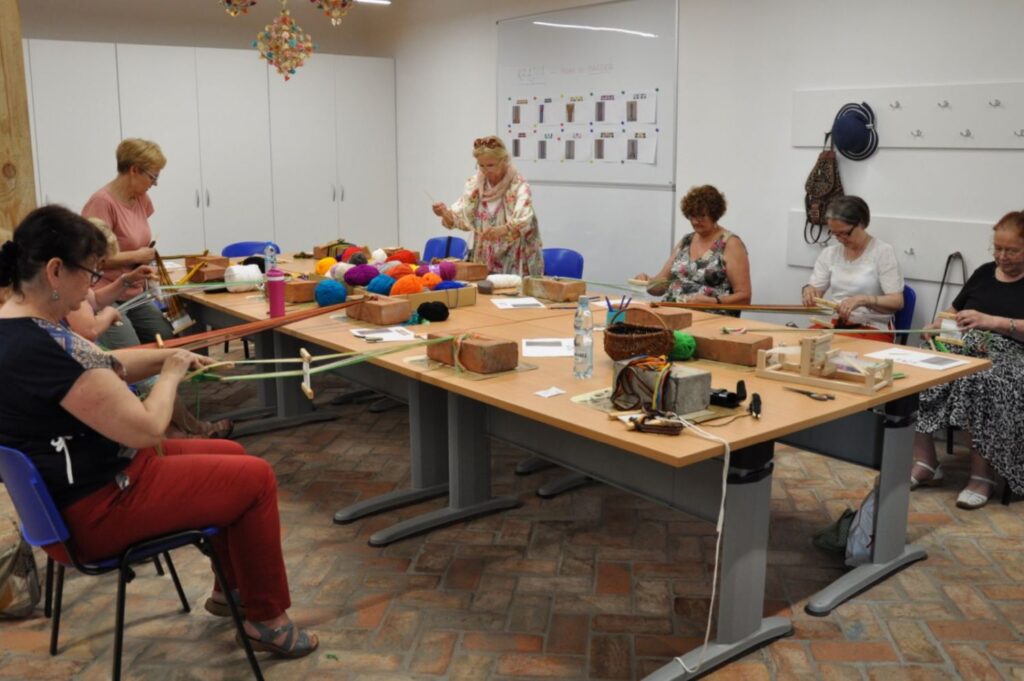 Uczestnicy warsztatów tkania na bardkach pracują przy stołach z kolorową włóczką i narzędziami tkackimi w sali warsztatowej (kliknięcie powiększy zdjęcie).