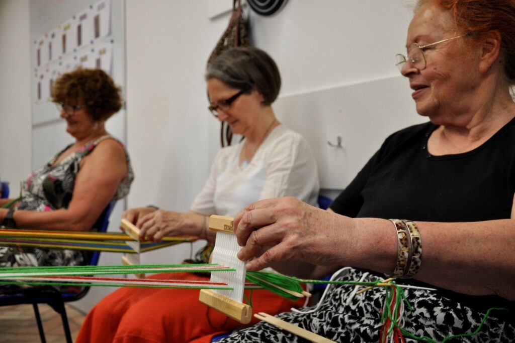 Trzy kobiety siedzące obok siebie tkają na bardkach, przeplatając barwne nici podczas warsztatów (kliknięcie powiększy zdjęcie).