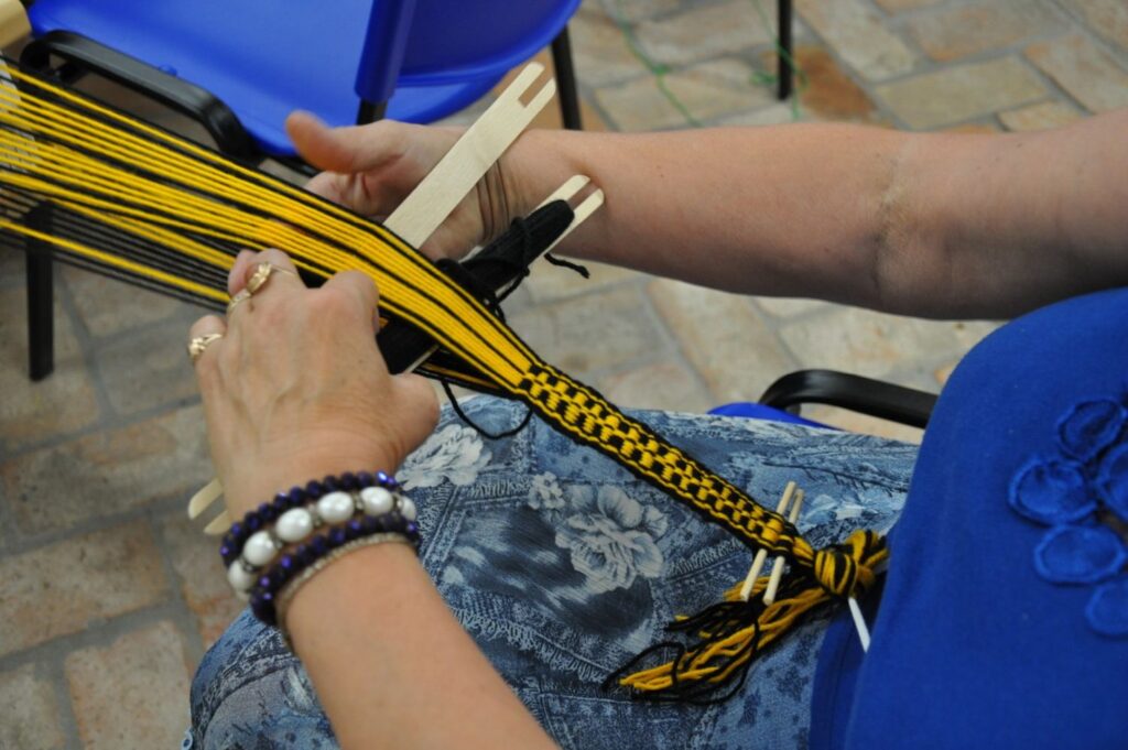 Uczestniczka warsztatów tkania na bardkach pracuje nad żółto-czarnym wzorem, trzymając napięte nici osnowy (kliknięcie powiększy zdjęcie).