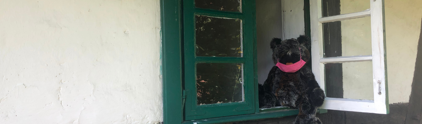 W otwartym oknie starej chałupy siedzi pluszowy misiu. Na pyszczku ma różową maseczkę ochronną.