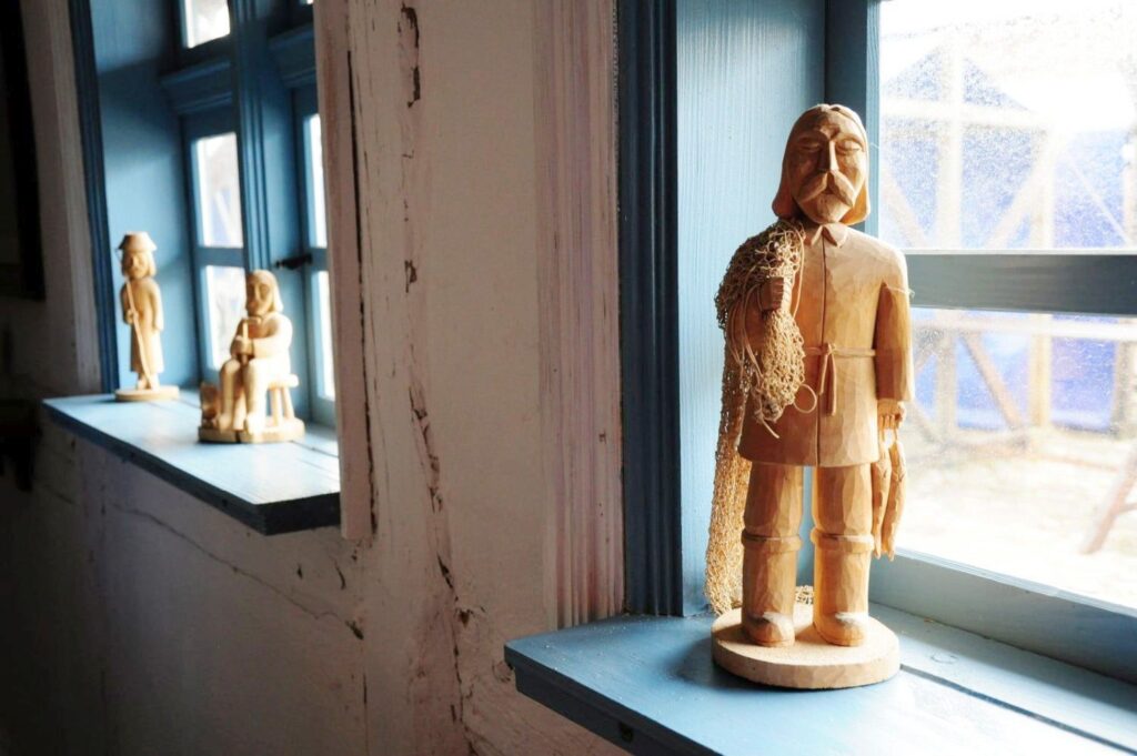 Drewniana rzeźba mężczyzny z siecią rybacką, stojąca na parapecie przy oknie, w tle widoczne mniejsze drewniane figurki ustawione w głębi pomieszczenia (kliknięcie powiększy zdjęcie).