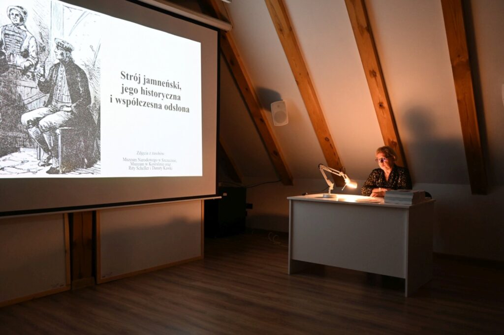 Prelegentka prowadzi wykład przy biurku, a na ekranie wyświetlona jest prezentacja o stroju jamneńskim (kliknięcie powiększy zdjęcie).