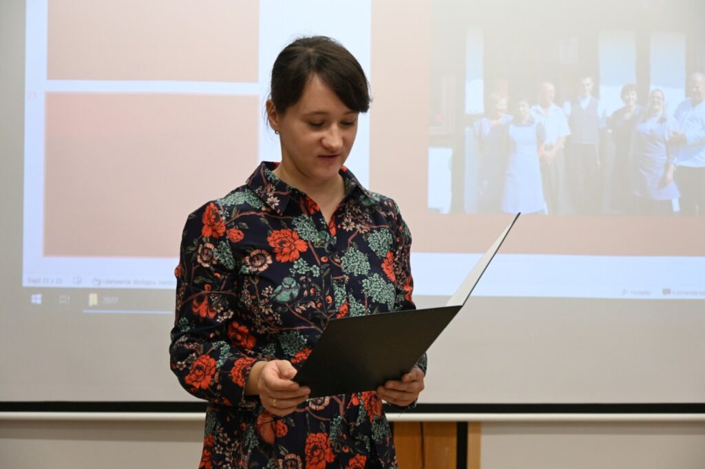 Kobieta w kwiecistej sukience stoi przed ekranem i czyta tekst podczas wystąpienia (kliknięcie powiększy zdjęcie)