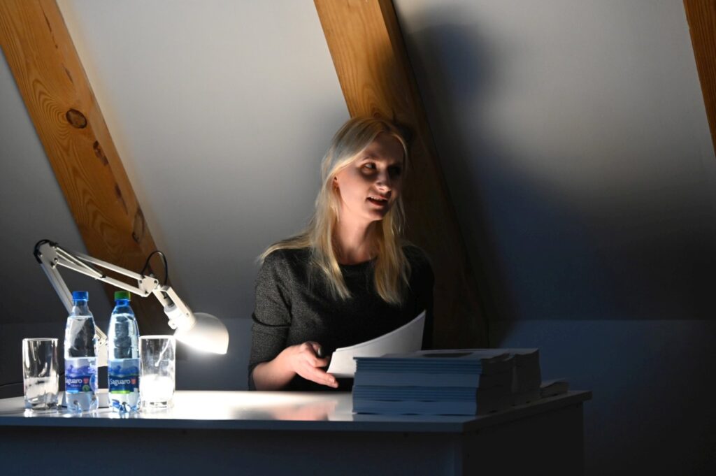 Prelegentka siedzi przy biurku z lampką i materiałami, prezentując temat podczas seminarium (kliknięcie powiększy zdjęcie).