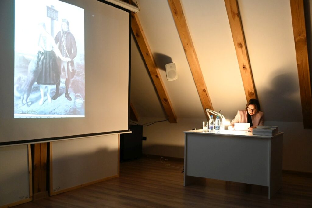 Wykład w sali poddasza – na ekranie wyświetlona archiwalna fotografia pary w strojach ludowych (kliknięcie powiększy zdjęcie).