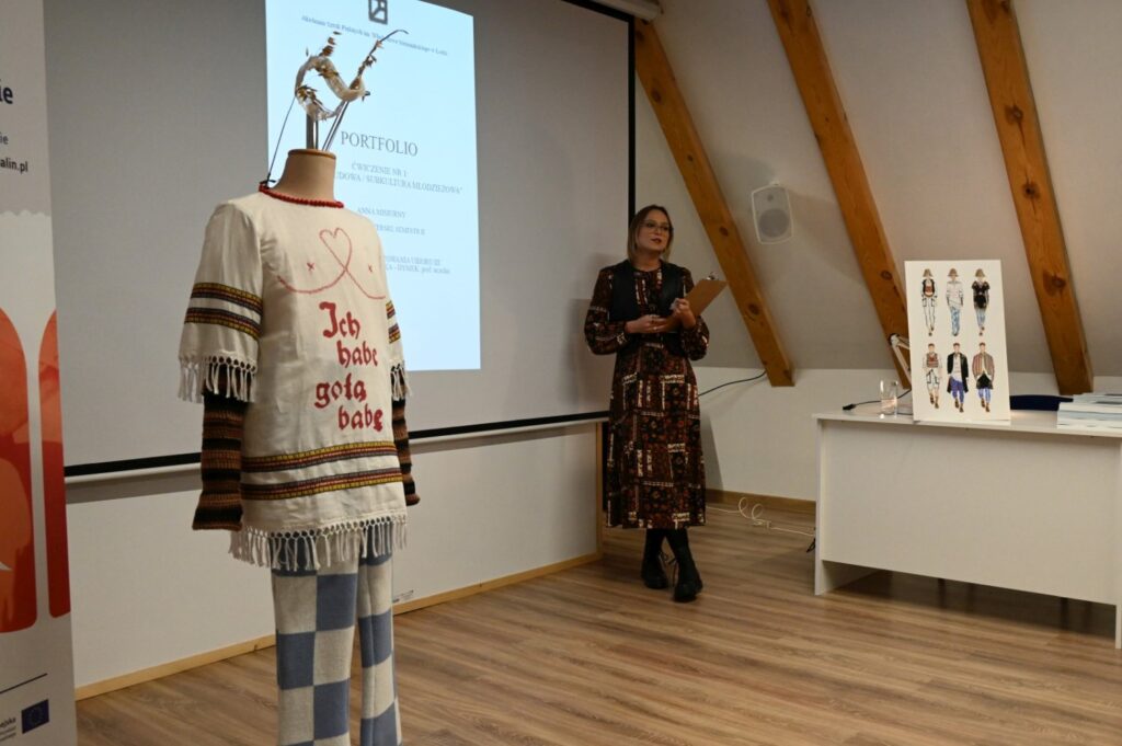 Prelegentka prowadzi wykład, po lewej stoi manekin ubrany w strój inspirowany strojem ludowym, obok plansze z ilustracjami ubiorów (kliknięcie powiększy zdjęcie).