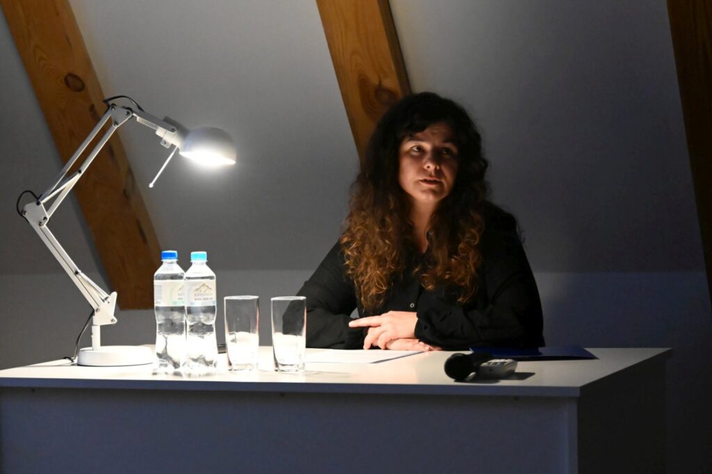 Kobieta z długimi włosami siedzi przy biurku oświetlonym lampką, przed nią stoją butelki wody i szklanki podczas wykładu (kliknięcie powiększy zdjęcie).