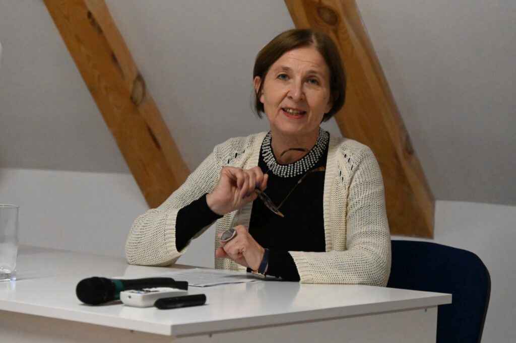 Kobieta siedzi przy biurku z mikrofonem, rozpoczynając swoje wystąpienie podczas seminarium (kliknięcie powiększy zdjęcie).