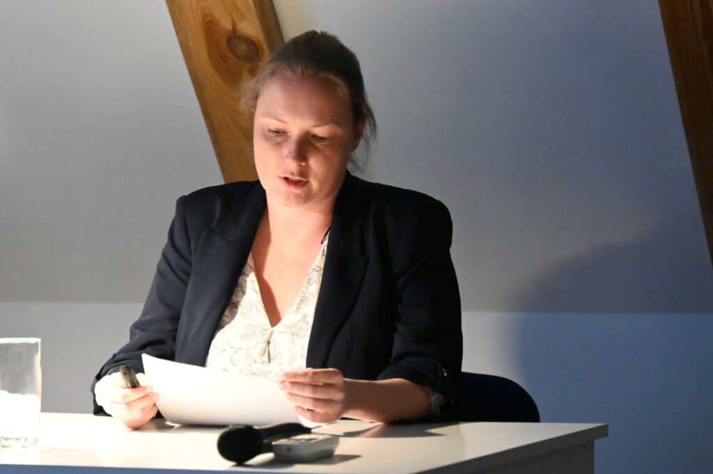 Prelegentka czyta z kartki przy oświetlonym biurku, prowadząc referat podczas spotkania (kliknięcie powiększy zdjęcie).