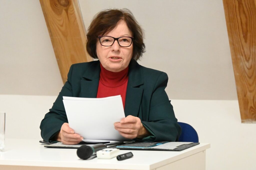 Kobieta w zielonej marynarce czyta przygotowany tekst przy biurku z mikrofonem, występując przed publicznością (kliknięcie powiększy zdjęcie).