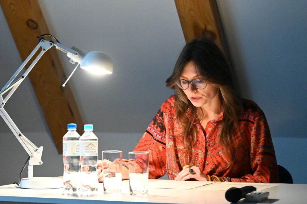 Prelegentka w czerwonej wzorzystej bluzce czyta notatki przy biurku z lampką i wodą, prowadząc wykład (kliknięcie powiększy zdjęcie).