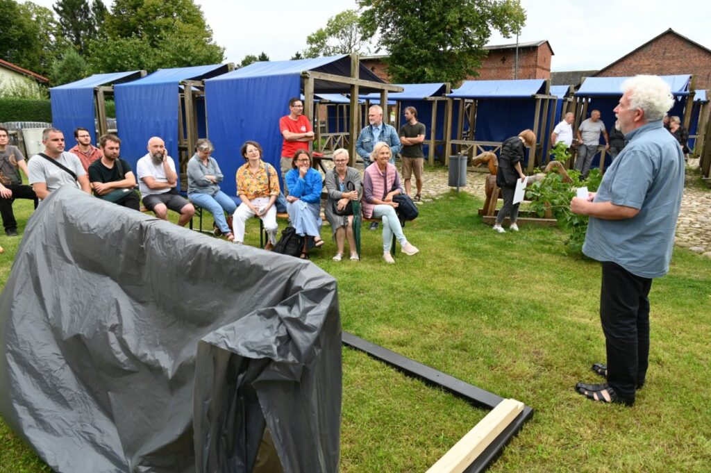Mężczyzna prowadzi spotkanie plenerowe na trawniku, stojąc przed siedzącą grupą uczestników, w tle widoczne są niebieskie namioty i zabudowania (kliknięcie powiększy zdjęcie).