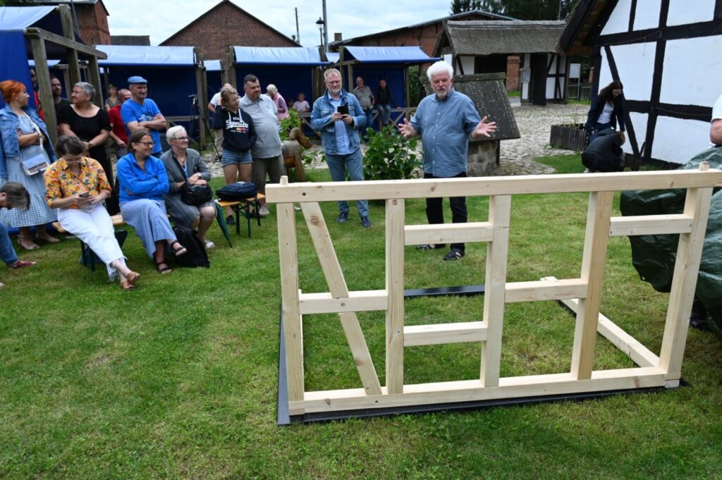 Uczestnicy wydarzenia stoją wokół drewnianej konstrukcji ściany szkieletowej, podczas gdy prowadzący omawia kolejne etapy montażu (kliknięcie powiększy zdjęcie).
