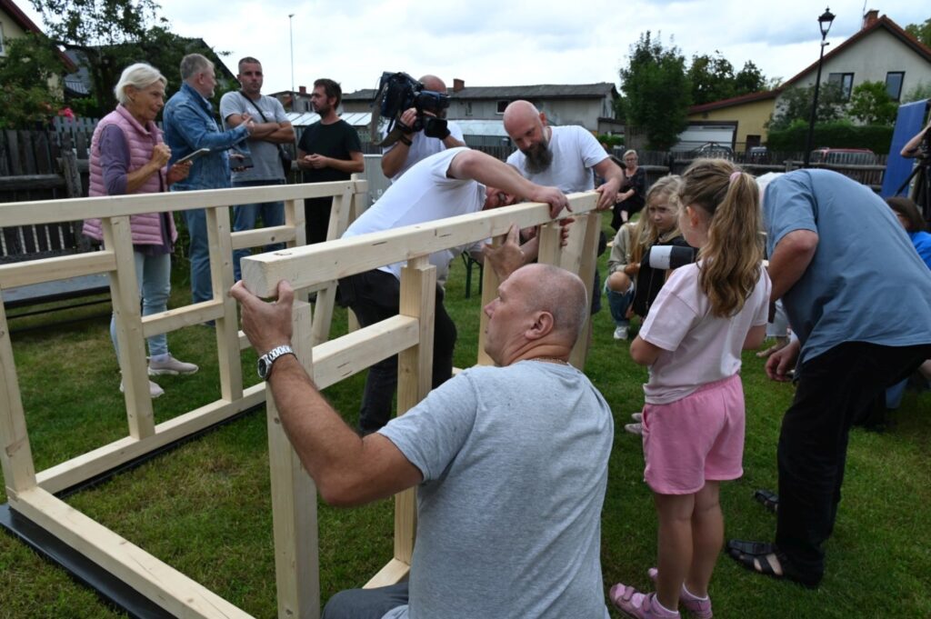 Uczestnicy wspólnie przykręcają kolejne elementy drewnianej ściany, pracując przy konstrukcji na trawniku (kliknięcie powiększy zdjęcie).