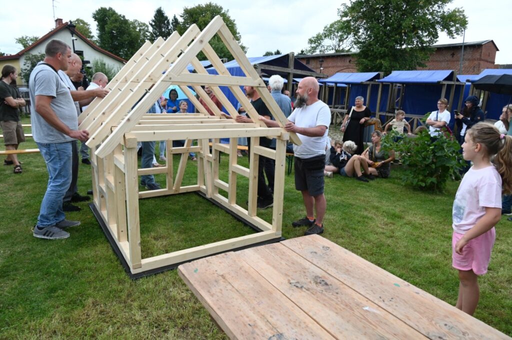 Grupa mężczyzn unosi i montuje drewnianą więźbę dachową na przygotowanej konstrukcji domu szkieletowego, wokół stoją uczestnicy wydarzenia (kliknięcie powiększy zdjęcie).