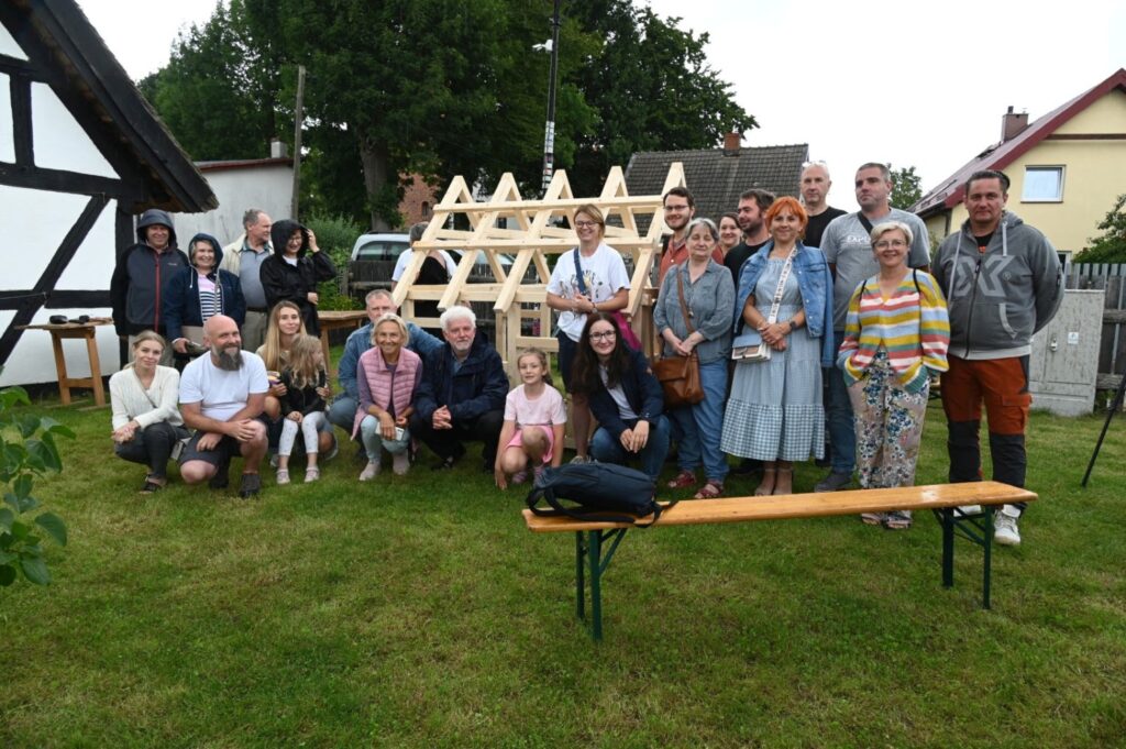 Uczestnicy warsztatów pozują do wspólnego zdjęcia przed zmontowaną drewnianą konstrukcją domu, siedząc i stojąc na trawie (kliknięcie powiększy zdjęcie).