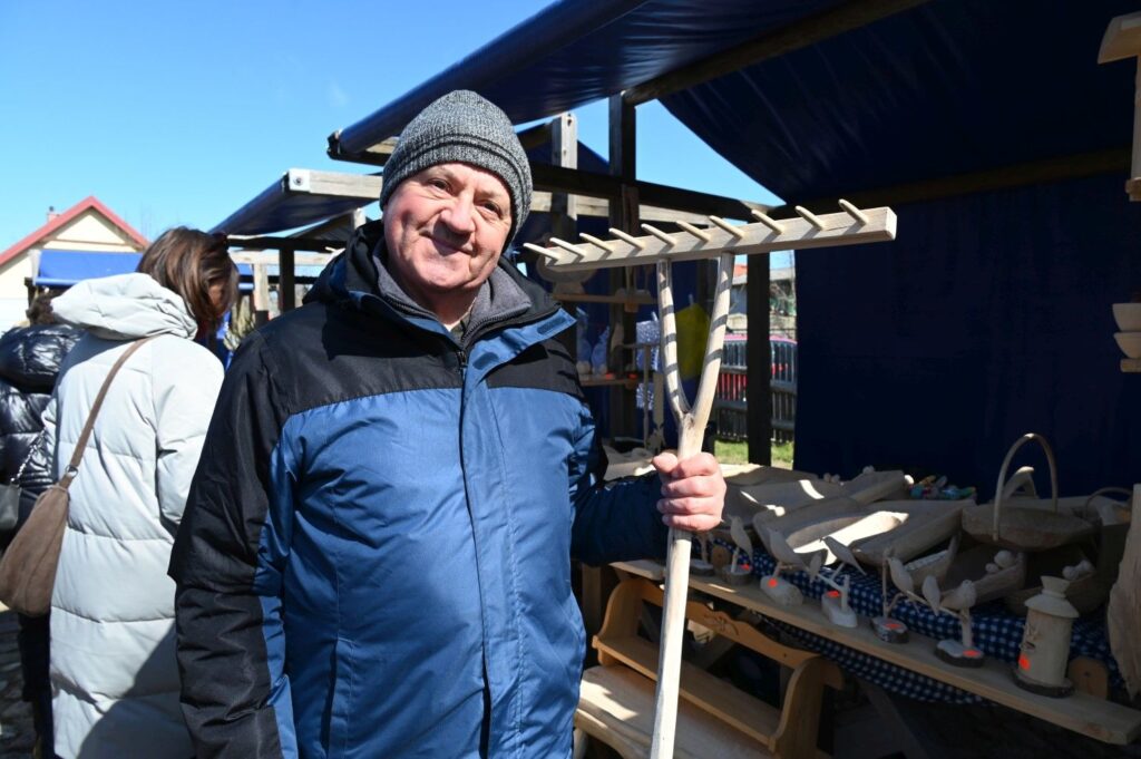 Mężczyzna stojący przy stoisku z rękodziełem na jarmarku wielkanocnym trzymający w dłoni drewniane grabie (kliknięcie powiększy zdjęcie).