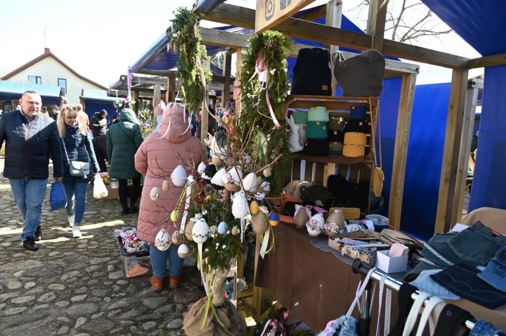 Stoisko z rękodziełem wielkanocnym ozdobione zielonymi gałązkami i pisankami, przy którym przechodzą odwiedzający jarmark (kliknięcie powiększy zdjęcie).