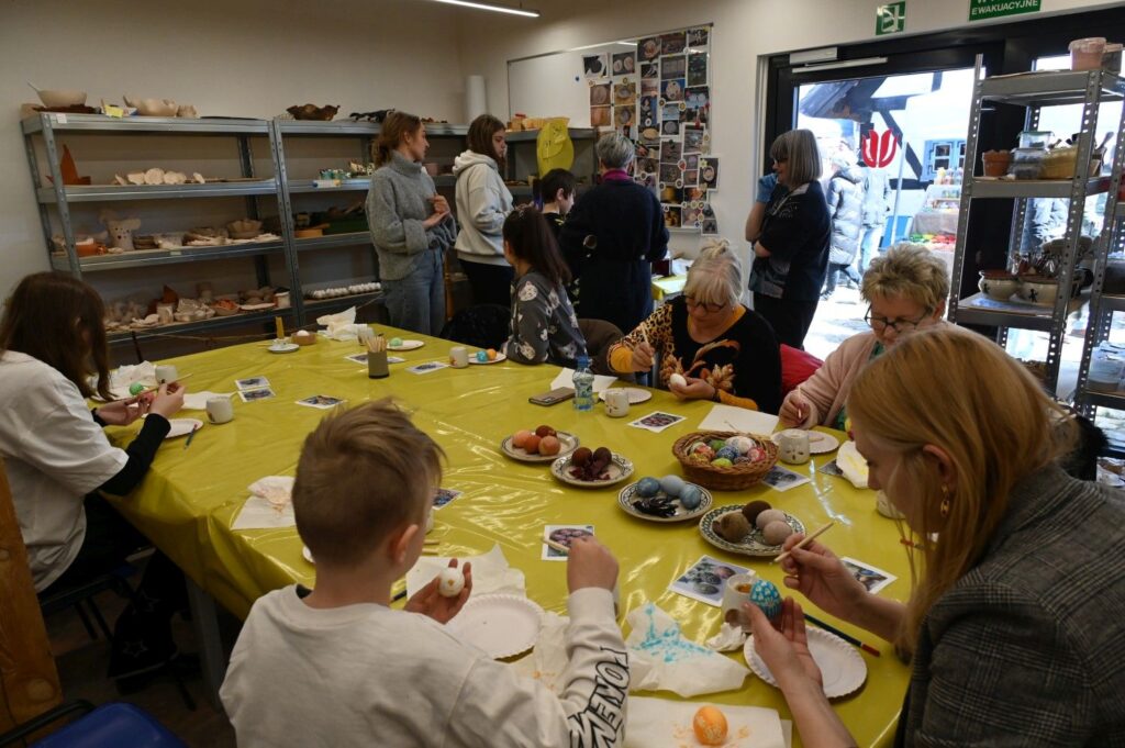 Uczestnicy warsztatów wielkanocnych siedzą przy stole i dekorują jajka podczas zajęć (kliknięcie powiększy zdjęcie).