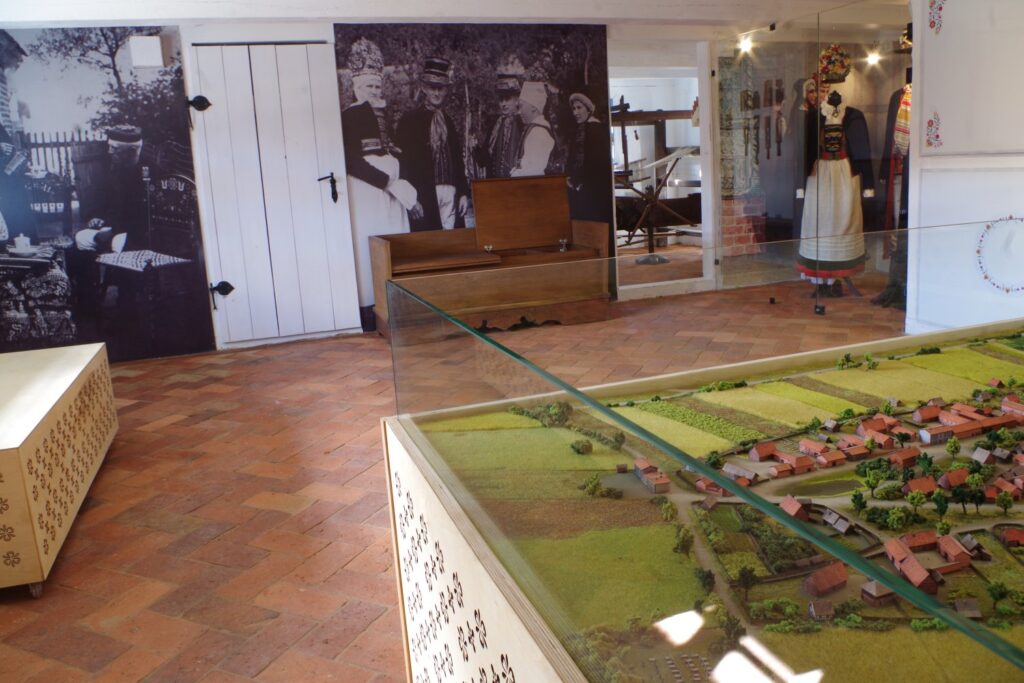 Wnętrze sali muzealnej z makietą wsi umieszczoną w gablocie oraz archiwalnymi fotografiami na ścianach (kliknięcie powiększy zdjęcie).