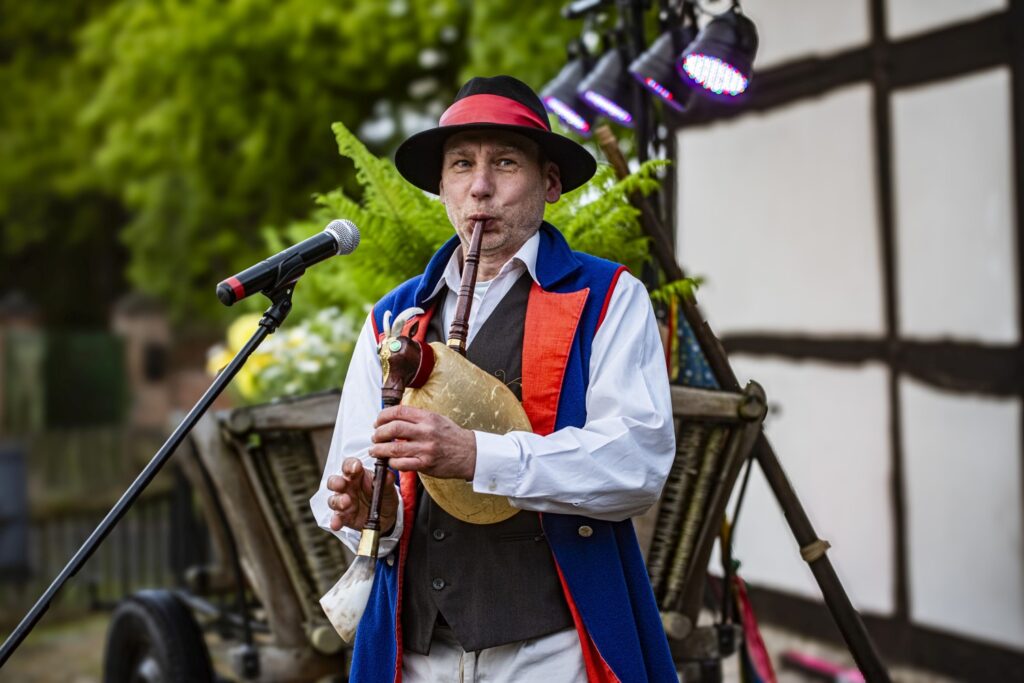 Muzyk w tradycyjnym stroju ludowym gra na dudach; występ odbywa się na tle zabudowy szachulcowej (kliknięcie powiększy zdjęcie).