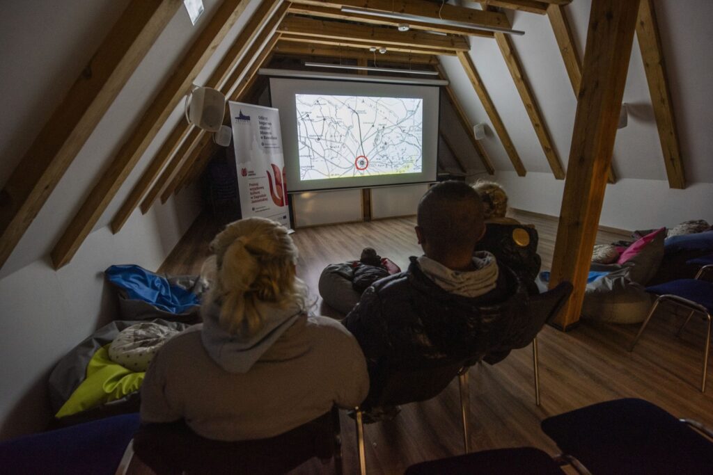Kilka osób siedzi w sali na poddaszu i ogląda prezentację multimedialną wyświetlaną na ekranie (kliknięcie powiększy zdjęcie).