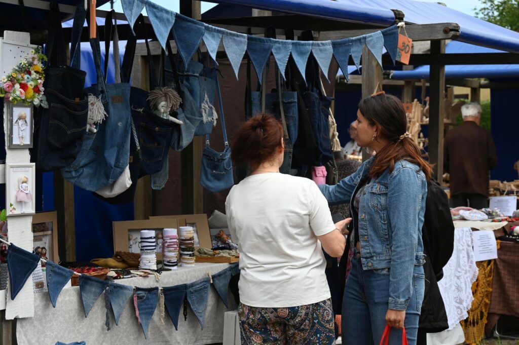 Dwie kobiety rozmawiające przy stoisku z torbami i wyrobami tekstylnymi na jarmarku (kliknięcie powiększy zdjęcie).
