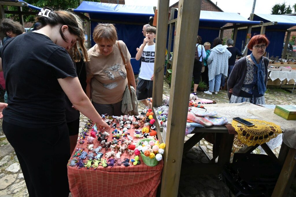 Odwiedzający przeglądający drobne kolorowe wyroby rękodzielnicze rozłożone na stole jarmarcznym (kliknięcie powiększy zdjęcie).