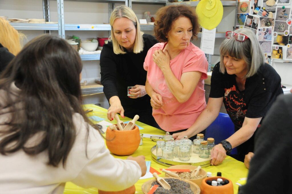 Uczestniczki warsztatów podczas zajęć, jedna osoba kładzie na stole deskę ze szklanymi słoikami (kliknięcie powiększy zdjęcie).