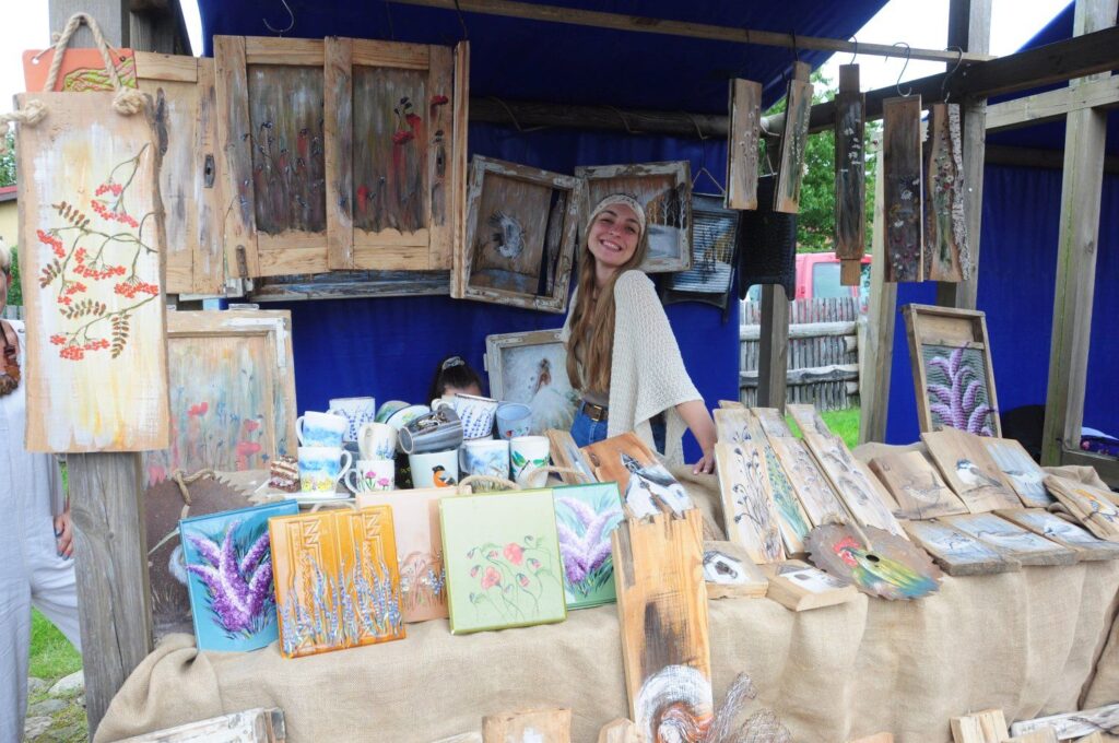 Stoisko z rękodziełem artystycznym, na którym wystawione są drewniane obrazy i drobne wyroby, przy stoisku stoi uśmiechnięta dziewczyna (kliknięcie powiększy zdjęcie).