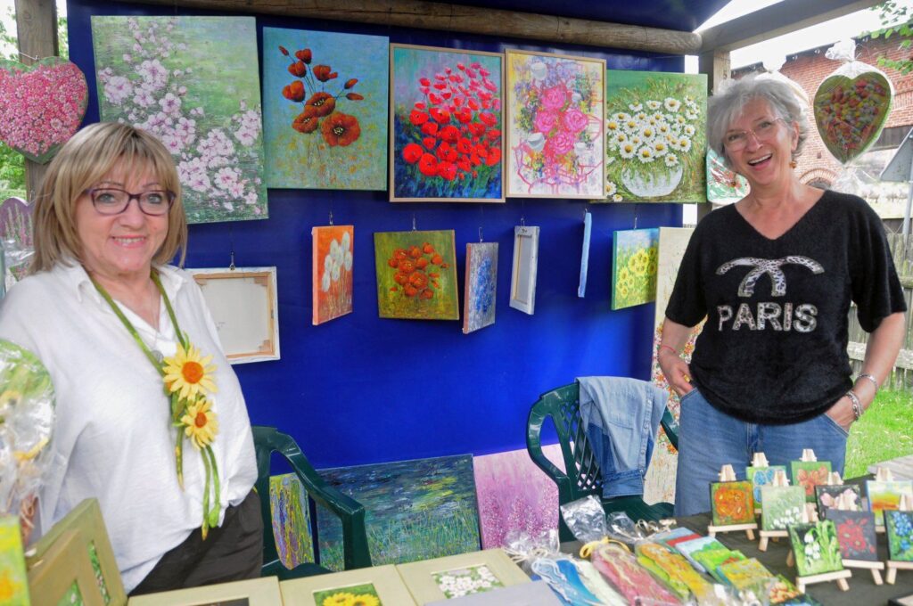 Dwie kobiety przy stoisku z obrazami i rękodziełem prezentują swoje prace podczas Jarmarku Jamneńskiego (kliknięcie powiększy zdjęcie).