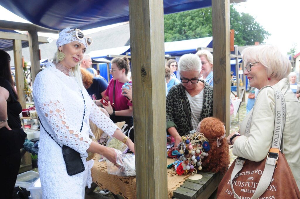 Stoisko z rękodziełem, przy którym odwiedzający oglądają ozdoby i rozmawiają z wystawczynią w białym stroju (kliknięcie powiększy zdjęcie).