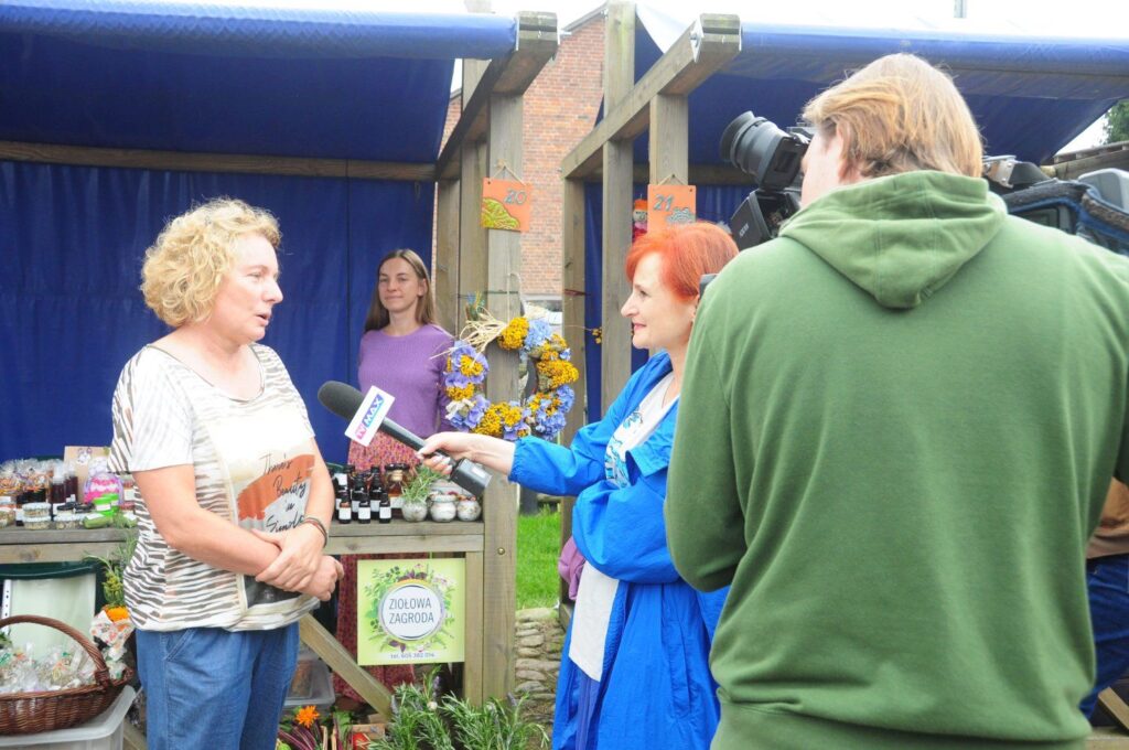 Reporterka przeprowadza wywiad z uczestniczką jarmarku, a obok stoi operator kamery nagrywający rozmowę (kliknięcie powiększy zdjęcie).