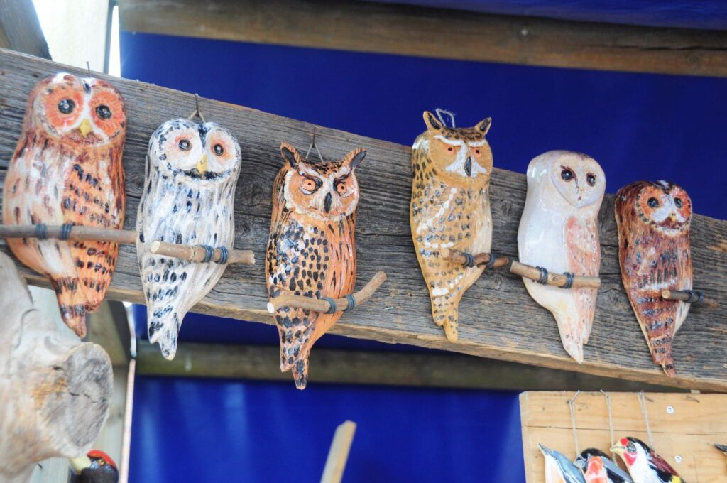 Ceramiczne figurki sów ustawione w rzędzie na drewnianej półce stoiska z rękodziełem (kliknięcie powiększy zdjęcie).