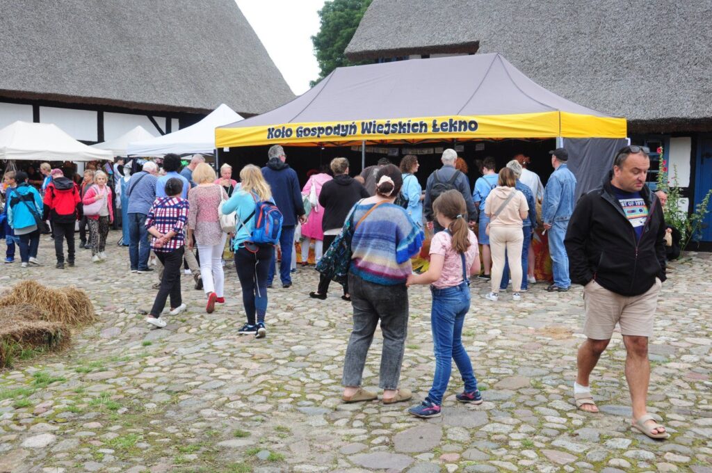 Tłum odwiedzających jarmark przed stoiskiem Koła Gospodyń Wiejskich, ustawionym przy tradycyjnej chałupie krytej strzechą (kliknięcie powiększy zdjęcie).