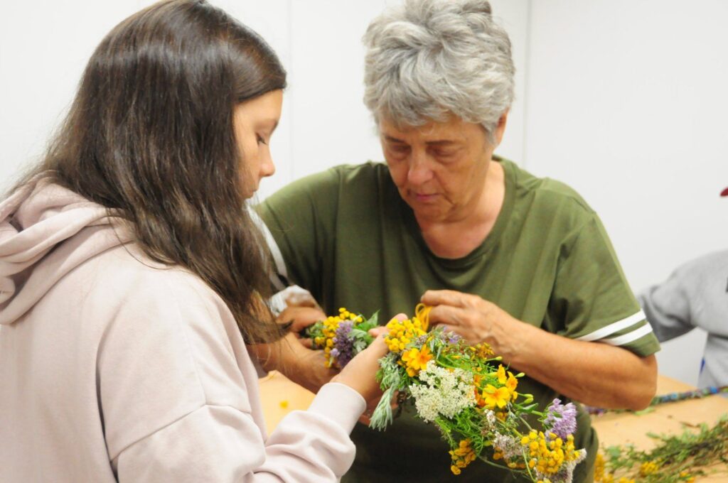 Uczestniczka warsztatów pomaga starszej kobiecie przy tworzeniu wianka z polnych kwiatów i ziół (kliknięcie powiększy zdjęcie).