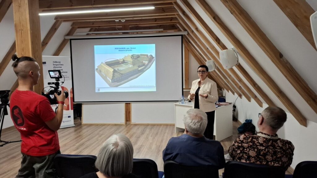 Prowadząca prezentująca slajd z modelem architektonicznym, uczestnicy siedzący na widowni, operator kamery nagrywający wydarzenie (kliknięcie powiększy zdjęcie)