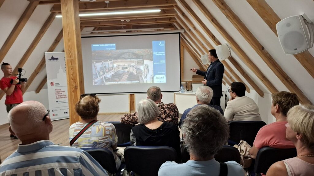 Uczestnicy wydarzenia siedzący na sali wykładowej, prelegent stojący przy ekranie z prezentacją architektoniczną (kliknięcie powiększy zdjęcie)