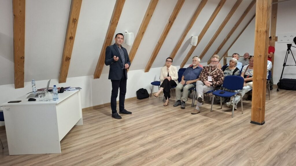 Prelegent stojący przed publicznością w sali z widoczną konstrukcją drewnianego dachu i stołem prezydialnym (kliknięcie powiększy zdjęcie)